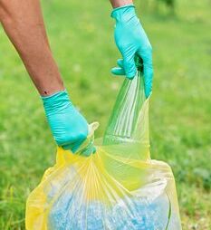 Person wearing gloves collecting recyclable bottles in a yellow plastic bag outdoors.